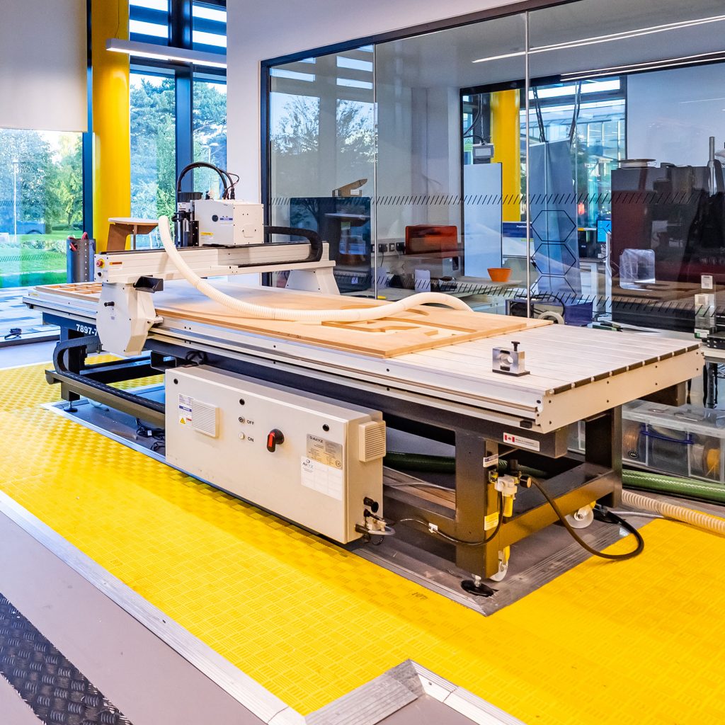 A CNC router in the Morson Engine Room, surrounded by yellow safety mats. Behind the router is glass that 