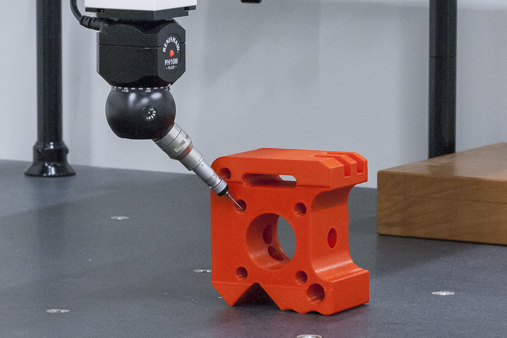 A CMM probe inspects holes on a red plastic 3D printed part.