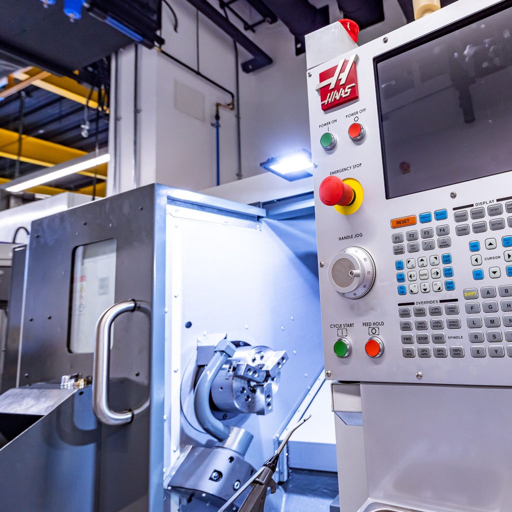 Looking into the open door of a lathe. A three jaw chuck is mounted to the spindle. On the right hand side of the image the Haas CNC control panel fills the frame.