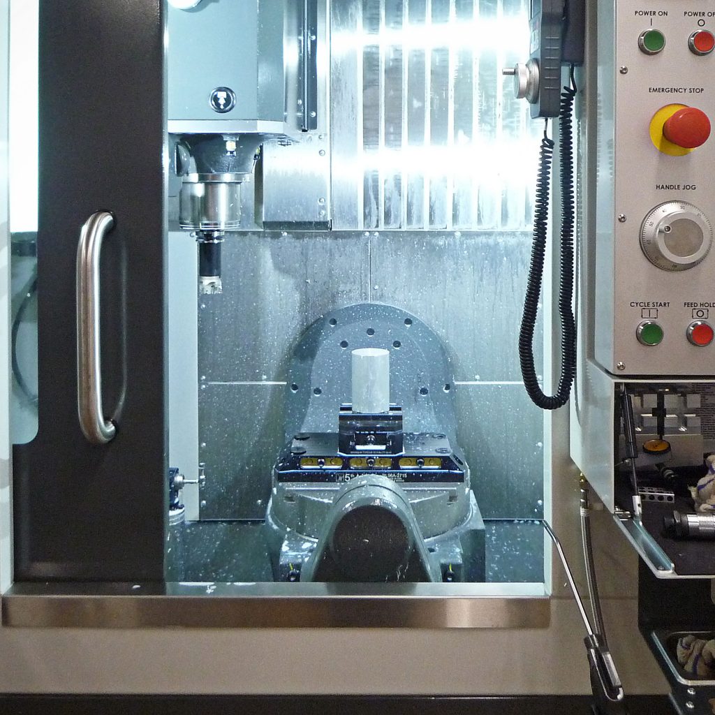 Looking through the open door of a Haas UMC-500 5-axis milling machine. In the vise centres on the circular machine table is a block of aluminium ready to be machined. There is a light coat of coolant coating the inside of the machine. In the spindle there is an inserted face mill.