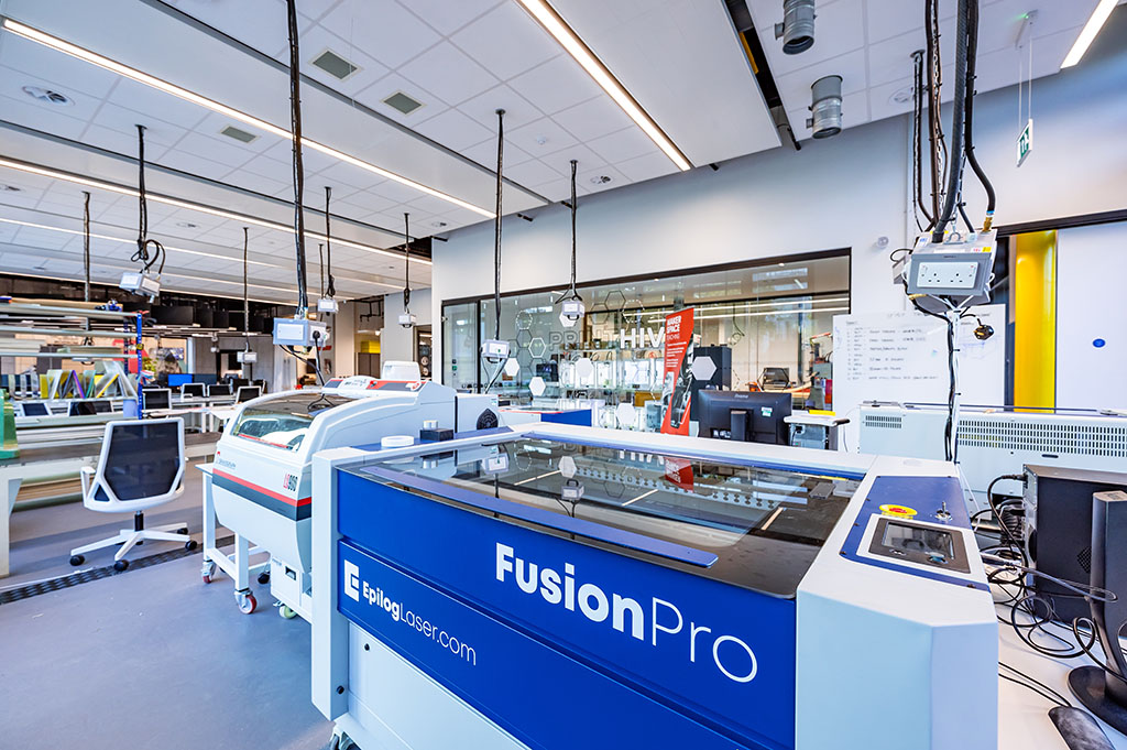 A workshop space with two laser cutters in the foreground. The main machine has Fusion Pro and EpilogLaser.com written on it. In the background is a glazed window with the words Print Hive on it. Throughout the workshop space power sockets hang from cables.