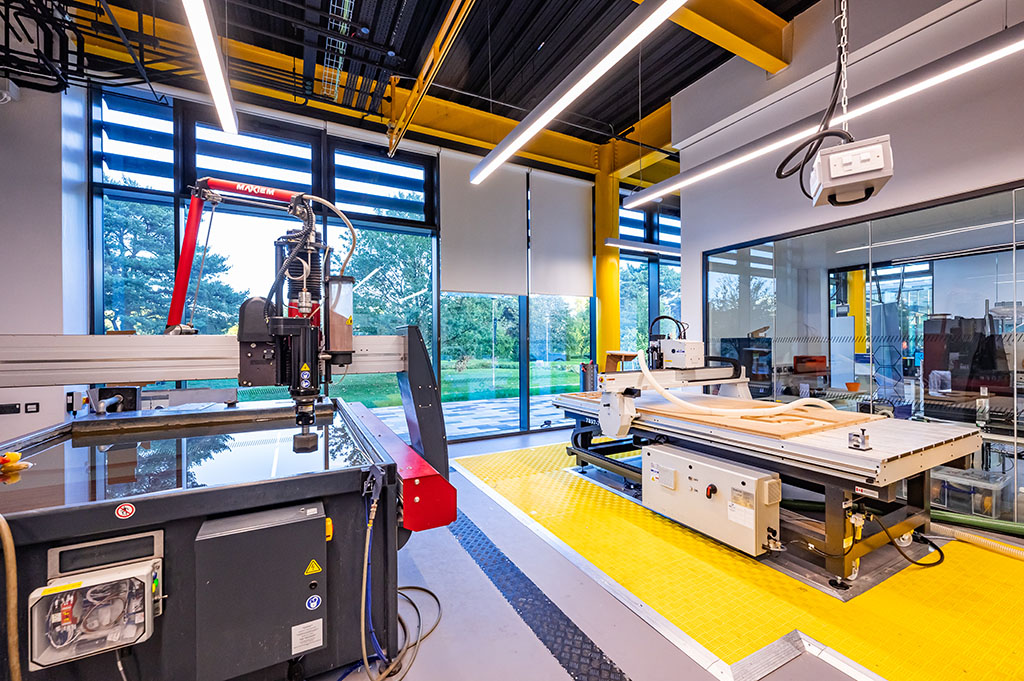 A waterjet cutter and a CNC router in the Morson Engine Room. The CNC router is surrounded by yellow safety mats. At the back of the image windows look out upon a paved path and trees.