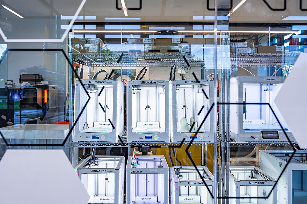 Ultimaker 3D printers sitting on metal wire rack shelves. There is a 3D print of a foot on one of the printers. In the foreground there is glass with vinyl hexagons.