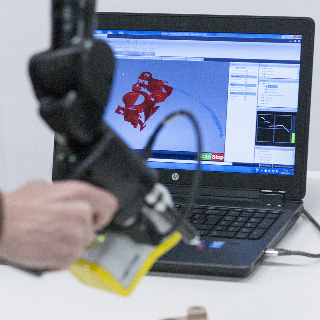 A hand in the foreground holds a laser 3D scanner. In the background a computer screen shows the result of the 3D scan data.