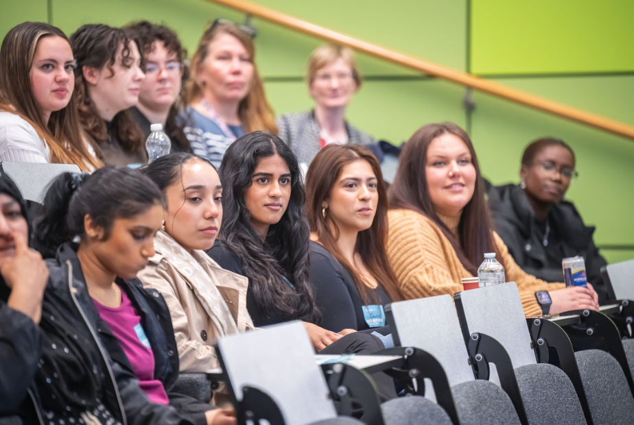 A large number of women sit in the rows of a lecture theatre. The wall behind them is a bright shade of green.