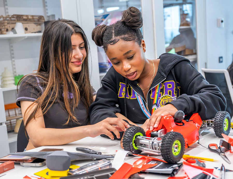 Two women working together to build a remote control racing car. The picture is colourful, with a red bodywork shell on the racing car. The woman on the left has long hair, and the woman on the right has her hair tied up.