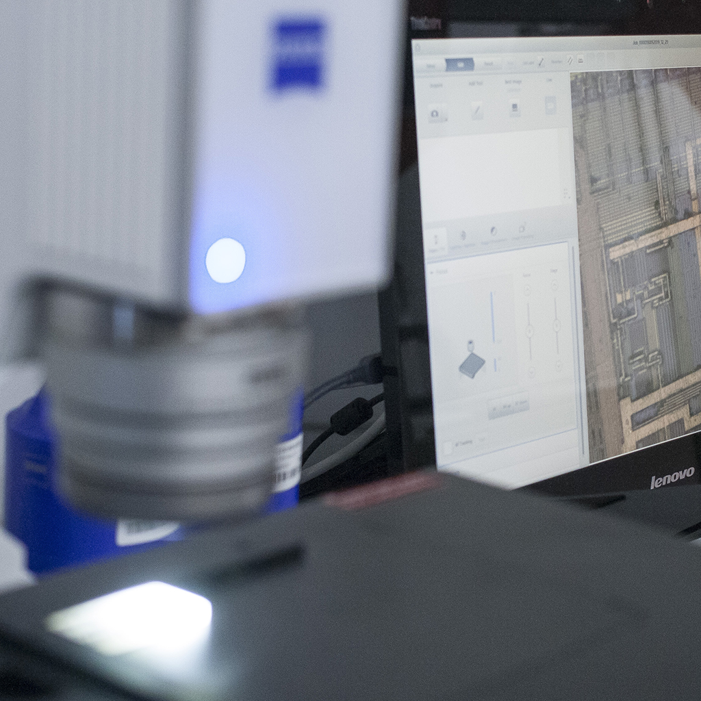 A digital microscope stage with a bright white light illuminating a sample. There is a blue LED light on the front of the microscope housing. In the background of the image a computer screen shows a microscope image.