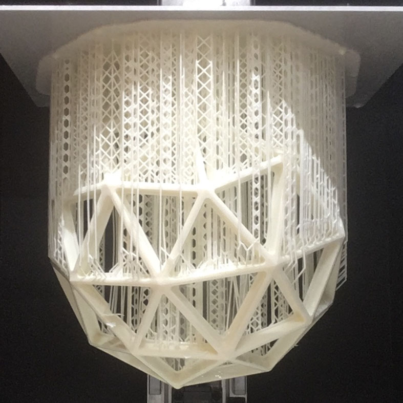 An off-white geodesic dome structure hangs suspended from the metal build plate of a resin 3D printer. The main structure is supported by lightweight, thin supports beams which can be snapped off later.