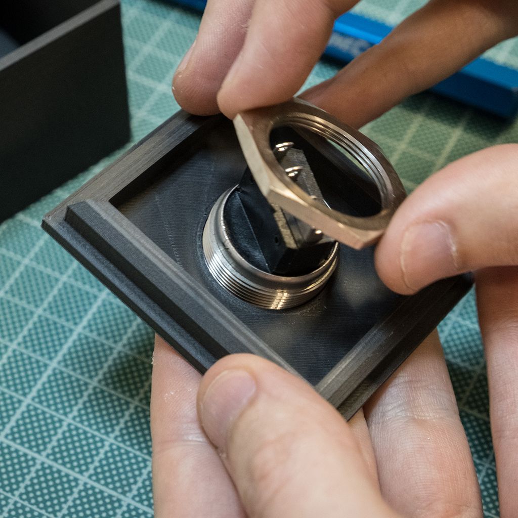 Fingers are twisting a threaded retaining ring onto the back of a switch. The switch has been inserted into a 3D printed component. Work is being performed on top of a green self-healing cutting mat with light grey grid marks on it. 