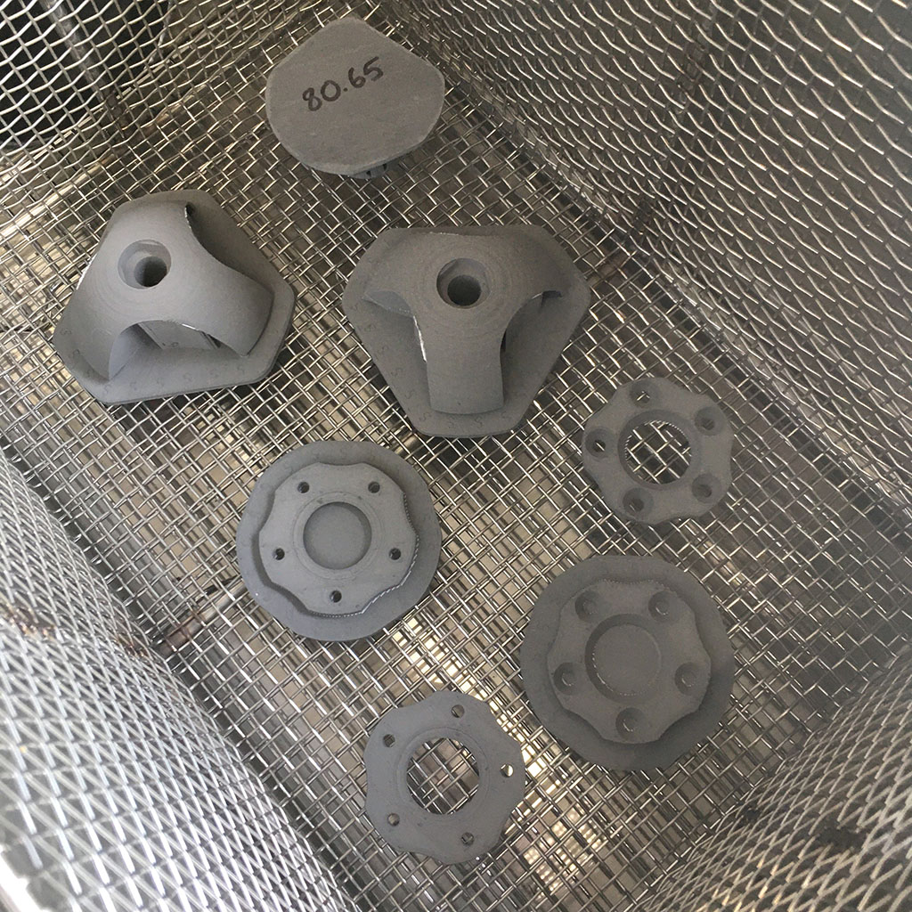 Metal 3D printed parts in a mesh basket waiting for sintering. The parts are a light grey. Two are ball clamps used for clamping pipe and tubing, the others are flat.