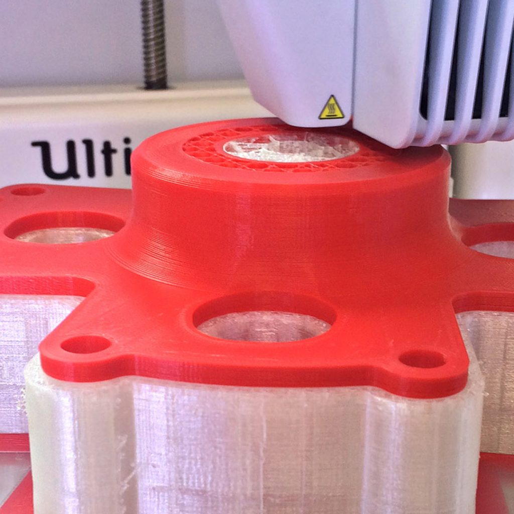 Red plastic 3D printer filament is being extruded into the form of a brake mount. Translucent support material holds up parts of the shape which are liable to sag. Behind the print the Ultimaker logo is partly shown. A yellow and black